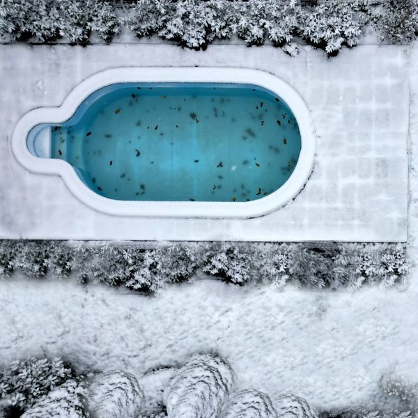An aerial view of a unique, keyhole-shaped inground swimming pool in winter. The surrounding deck and landscaping are covered in fresh white snow. The pool water is still visible, appearing light blue with some scattered leaves floating on its surface. An aerial view of a unique, keyhole-shaped inground swimming pool in winter. The surrounding deck and landscaping are covered in fresh white snow. The pool water is still visible, appearing light blue with some scattered leaves floating on its surface.