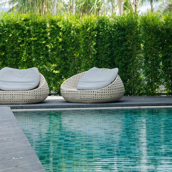 Two modern, light-colored, woven outdoor lounge chairs with white cushions sitting on a dark grey poolside patio next to a swimming pool with light blue and green mosaic tiles. A lush green hedge forms the background. Two modern, light-colored, woven outdoor lounge chairs with white cushions sitting on a dark grey poolside patio next to a swimming pool with light blue and green mosaic tiles. A lush green hedge forms the background.
