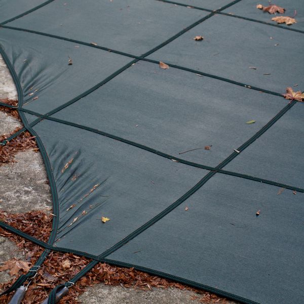A close-up view of a dark green mesh winter pool cover, secured by black straps, lying on a concrete patio. Scattered brown autumn leaves are visible on and around the cover, indicating the season. A close-up view of a dark green mesh winter pool cover, secured by black straps, lying on a concrete patio. Scattered brown autumn leaves are visible on and around the cover, indicating the season.