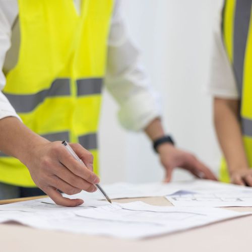 Person in yellow vest reviewing blueprints with colleague.