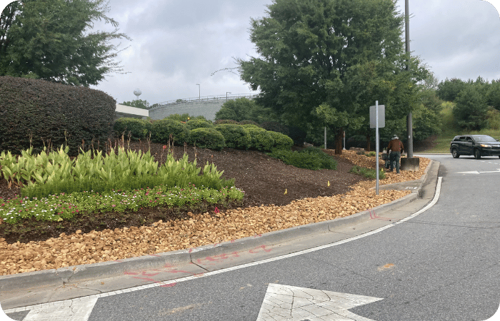 Curved garden bed with plants and rocks along a roadside sidewalk.