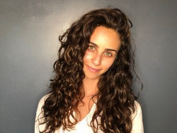Woman with long, curly brown hair wearing a white top standing in front of a gray wall at THE LONDONER in Hermosa Beach.