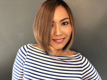 Woman with a striped black and white shirt and styled brown hair standing in front of a gray background at THE LONDONER's Hermosa Beach hair salon.