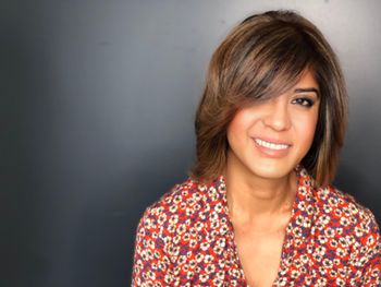 Woman wearing a white and red floral shirt standing in front of a gray wall inside of THE LONDONER's Hermosa Beach hair salon.