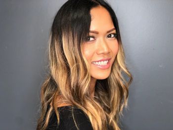 Woman with brown, highlighted hair smiling and standing in front of a gray background at THE LONDONER in Hermosa Beach.