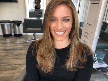 Woman with long brunette hair wearing a black top and standing inside of THE LONDONER's Hermosa Beach hair salon.