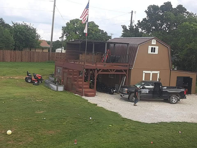 A small brown barn featuring a metal roof and an elevated wooden deck flying the American and Texas flags.