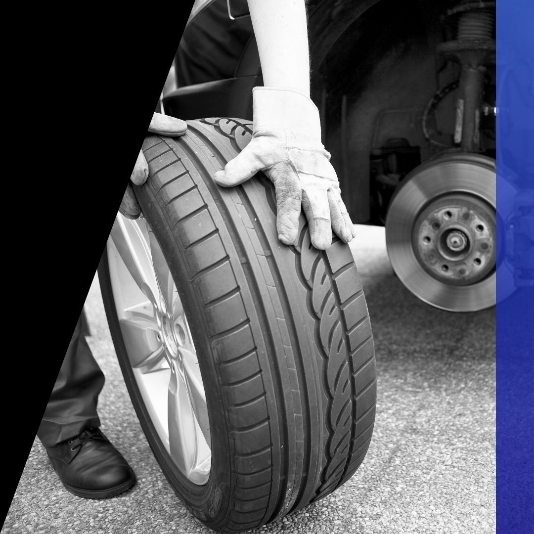 Close-up of a gloved hand rolling a passenger vehicle tire with exposed brake rotors in the background, representing car tire and brake services.