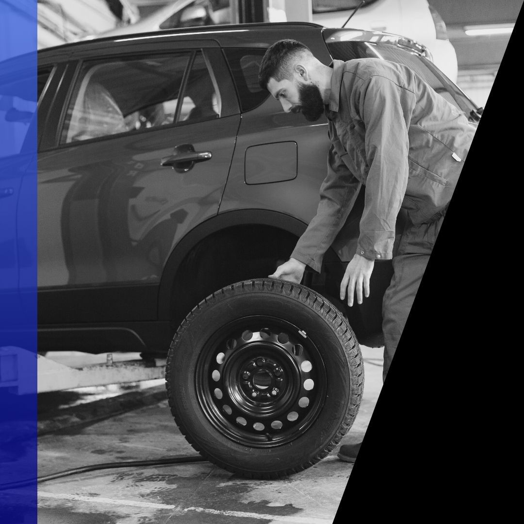 Male mechanic placing a new tire next to a passenger SUV raised on a lift in the shop, illustrating in-shop tire service.