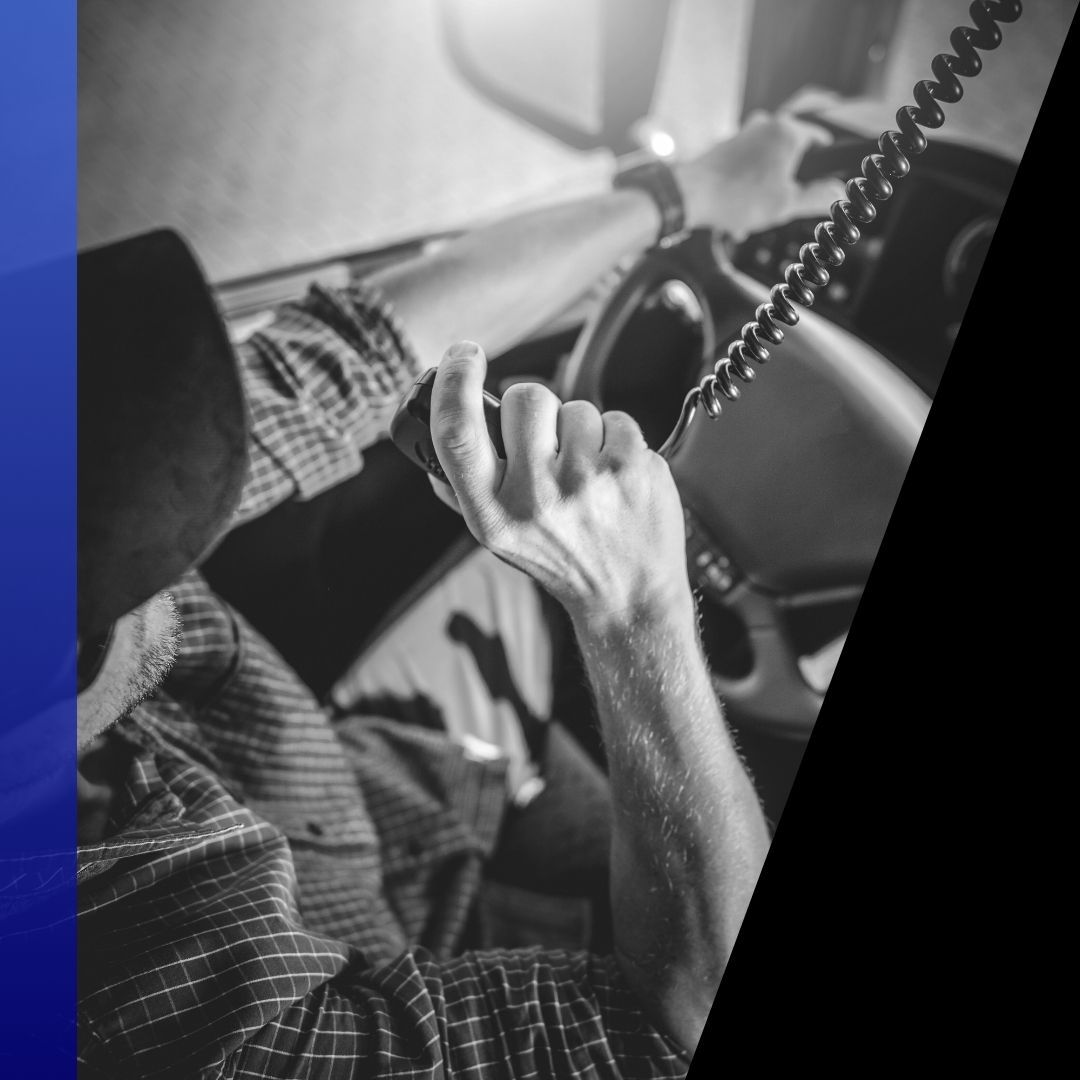 Overhead view of a truck driver's hands holding a CB radio microphone inside the semi-truck cab, symbolizing communication and roadside assistance.