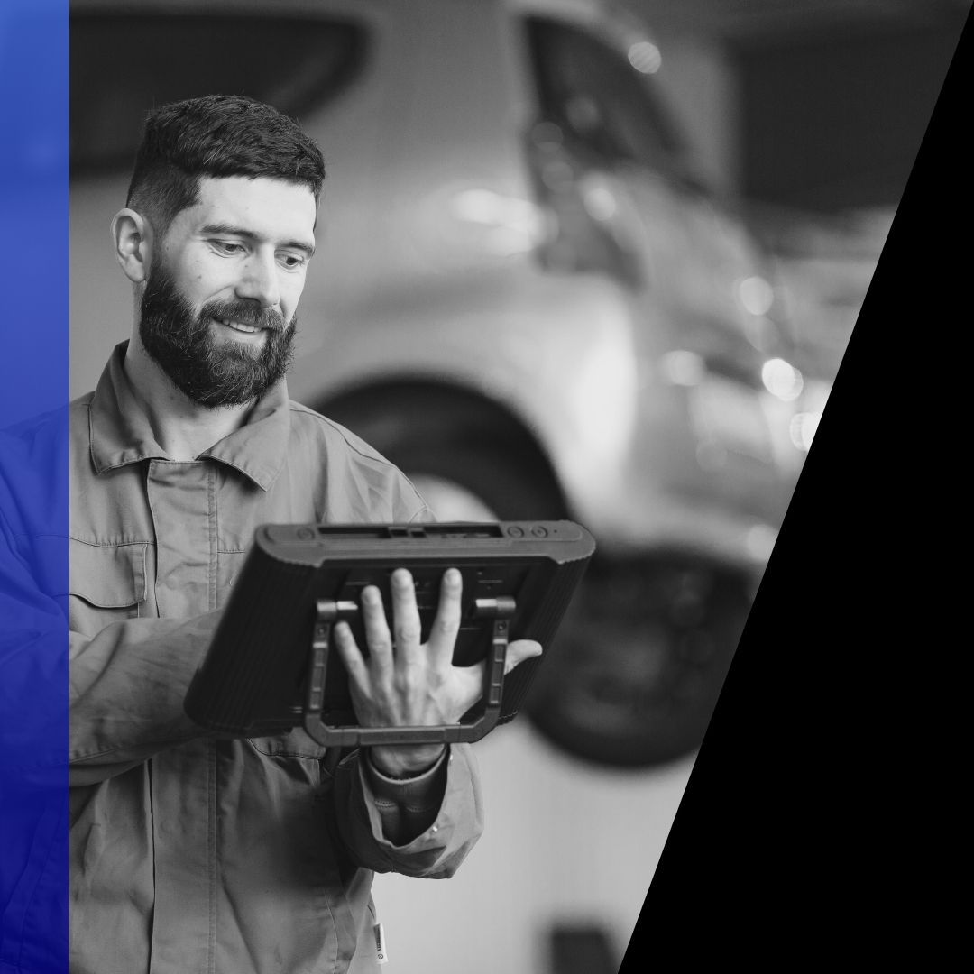 Male mechanic with a beard smiling while reading diagnostic data on a tablet inside the shop, conveying professional service.