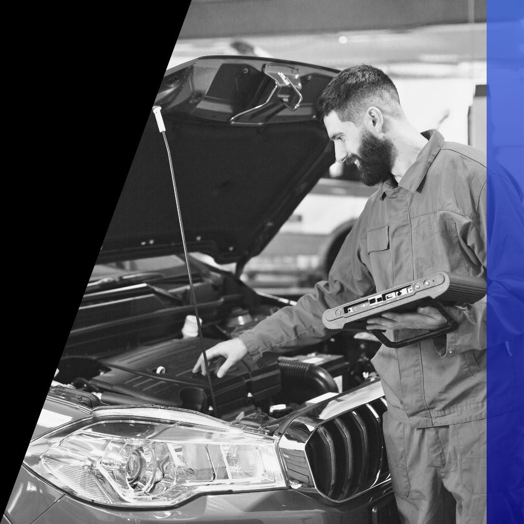 Male mechanic with a beard smiling while holding a diagnostic tablet and checking the engine bay of a passenger car in a shop.