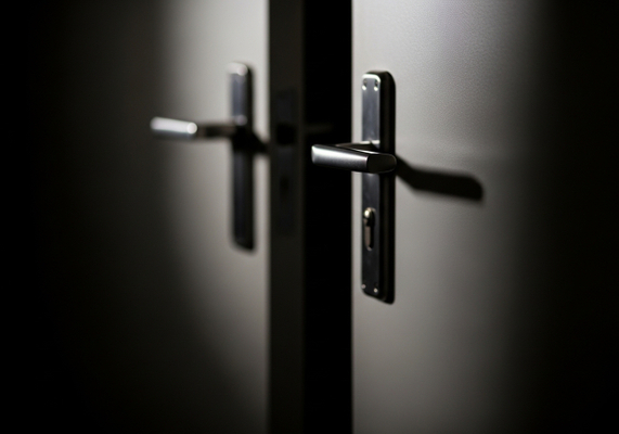 A close-up shot of a closed door featuring a modern, brushed metal door handle. The door appears to be gray or a similar neutral tone, with a smooth texture. The lighting is dramatic, casting a sharp shadow from the handle onto the door, creating a sense of depth and highlighting the door's surface. The focus is on the handle and its immediate surroundings, with the rest of the door fading into darkness, adding an element of mystery or intrigue. Closed door with handle