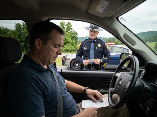 A man in a car looks at documents while a uniformed police officer stands outside the driver's window holding an ID card. A police car is in the background. Driver and Police Officer During a Traffic Stop