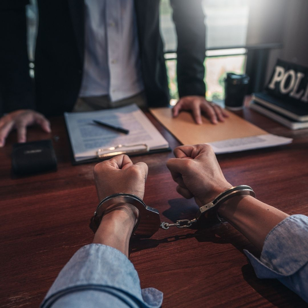 Person in handcuffs sits across a desk from an officer in an interrogation room.