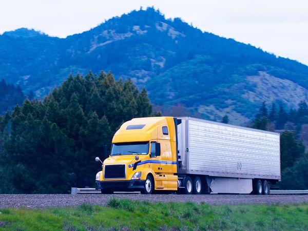 Modern popular powerful yellow big rig semi truck with a refrigerator trailer transports frozen food products from California to Oregon in the scenic highway among mountain passes rising above the distance.