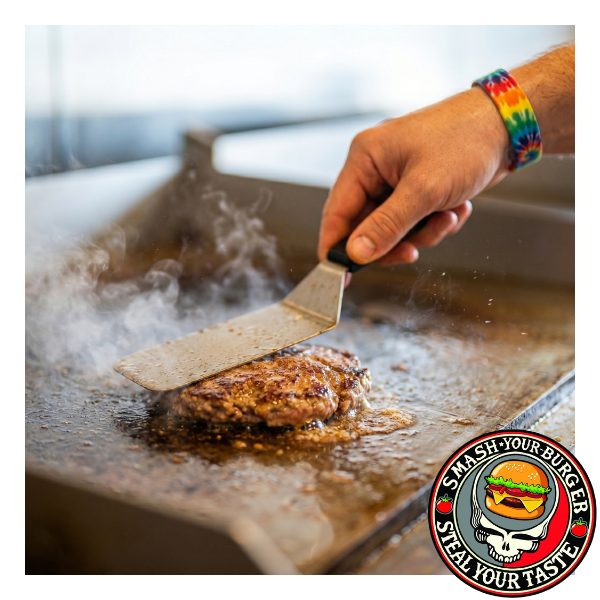 A close-up 1:1 photograph focused on a chef's hand with a tie-dye wristband pressing down on a sizzling beef burger patty with a spatula on a hot steel griddle, capturing motion and steam.