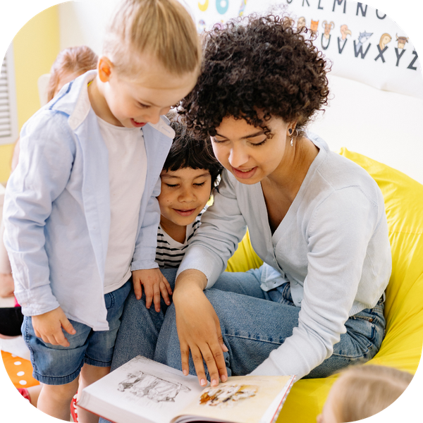 a woman sitting in a classroom with 2 young children close to her as she reads from a book 