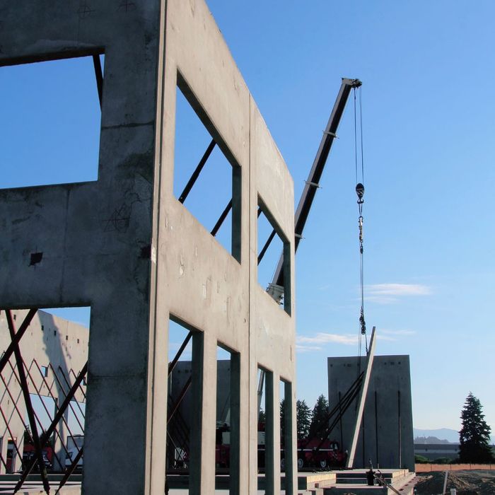A stock-style image of a precast concrete wall panel being lifted into place by a crane at a commercial construction site.