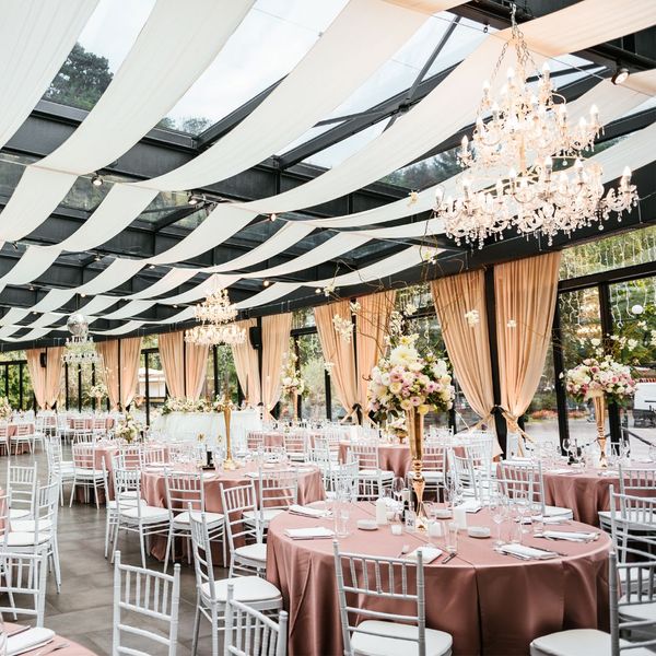 a large wedding venue decorated with rose pink tables with tall flowers, chandeliers, and ceiling and wall drapes