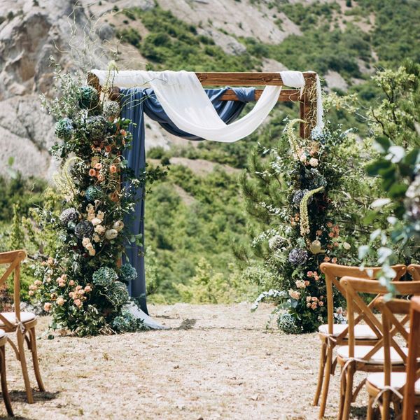 a wedding arch decorated in large dark foliage and navy blue and white drapes in front of a mountain