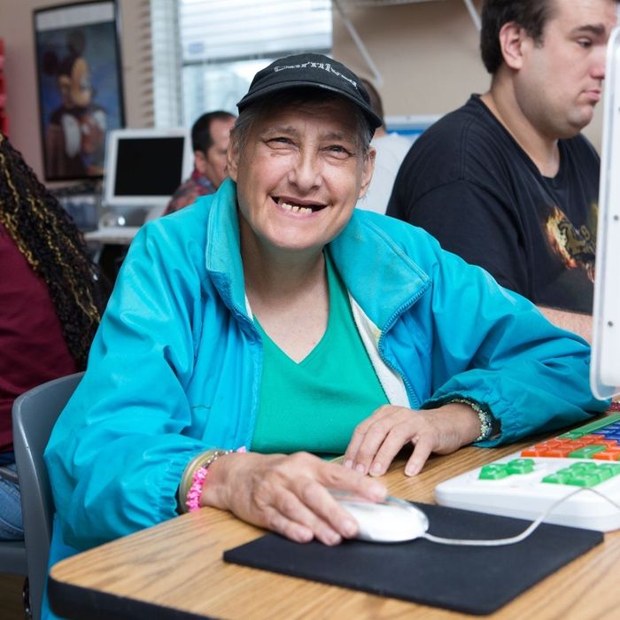 A smiling female participant wearing a blue jacket and a baseball cap is focused on using a computer mouse and keyboard during a learning or activity session at a UCO center.