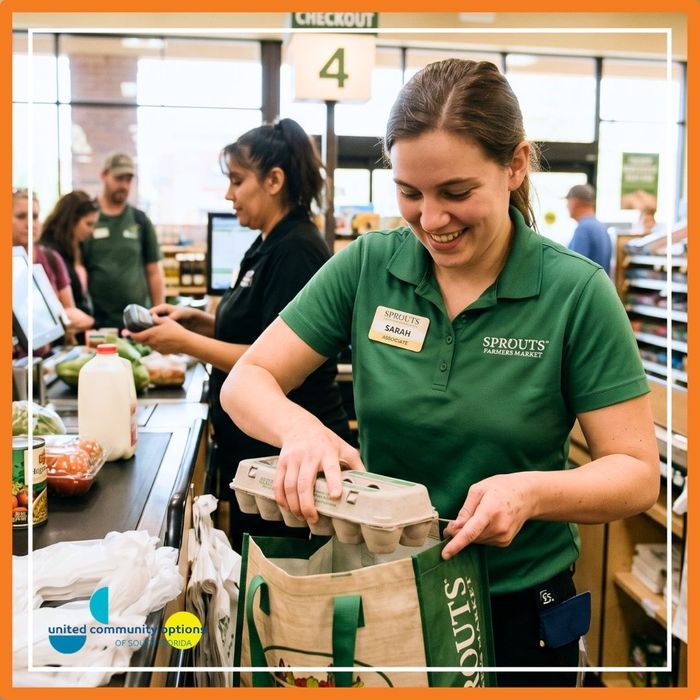 A retail associate in a green uniform smiling while carefully bagging groceries at a supermarket checkout station.