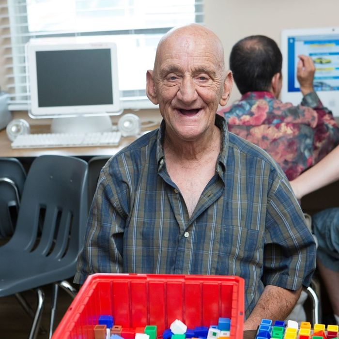 A happy older male participant with a warm, toothy smile sits at a computer station with a box of colorful blocks in the foreground, showcasing engagement at a UCO center.