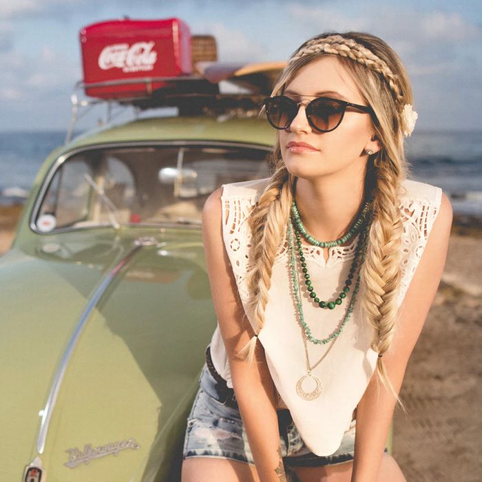 Girl with two fishtail braids sits on old, green VW bug parked on beach.