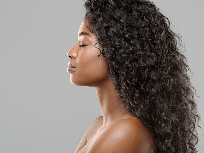 Profile portrait of beautiful African American woman with long curly hair extensions against a light gray background.