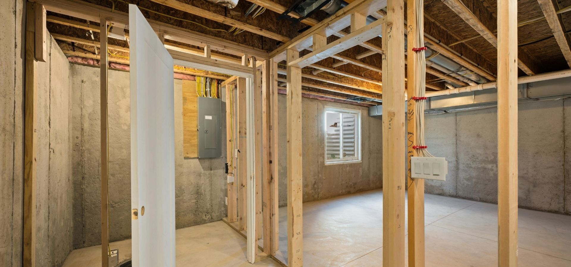 Interior view of a new construction home in the framing stage, showing a concrete basement foundation, timber wall studs, and professional electrical panel installation.
