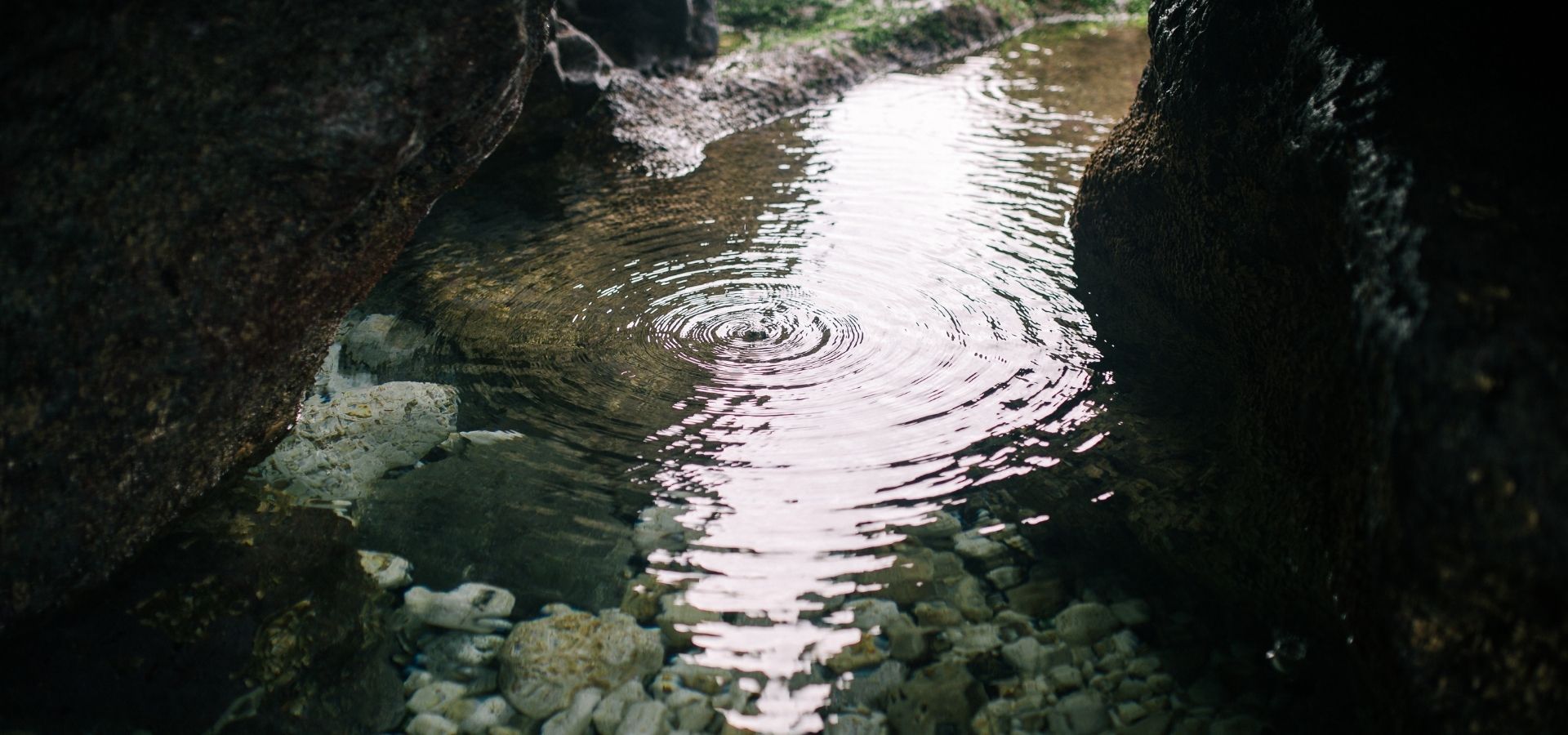 An artistic close-up of a serene garden pond with ripples in the water, natural rock borders, and clear visibility of the pebble bottom.