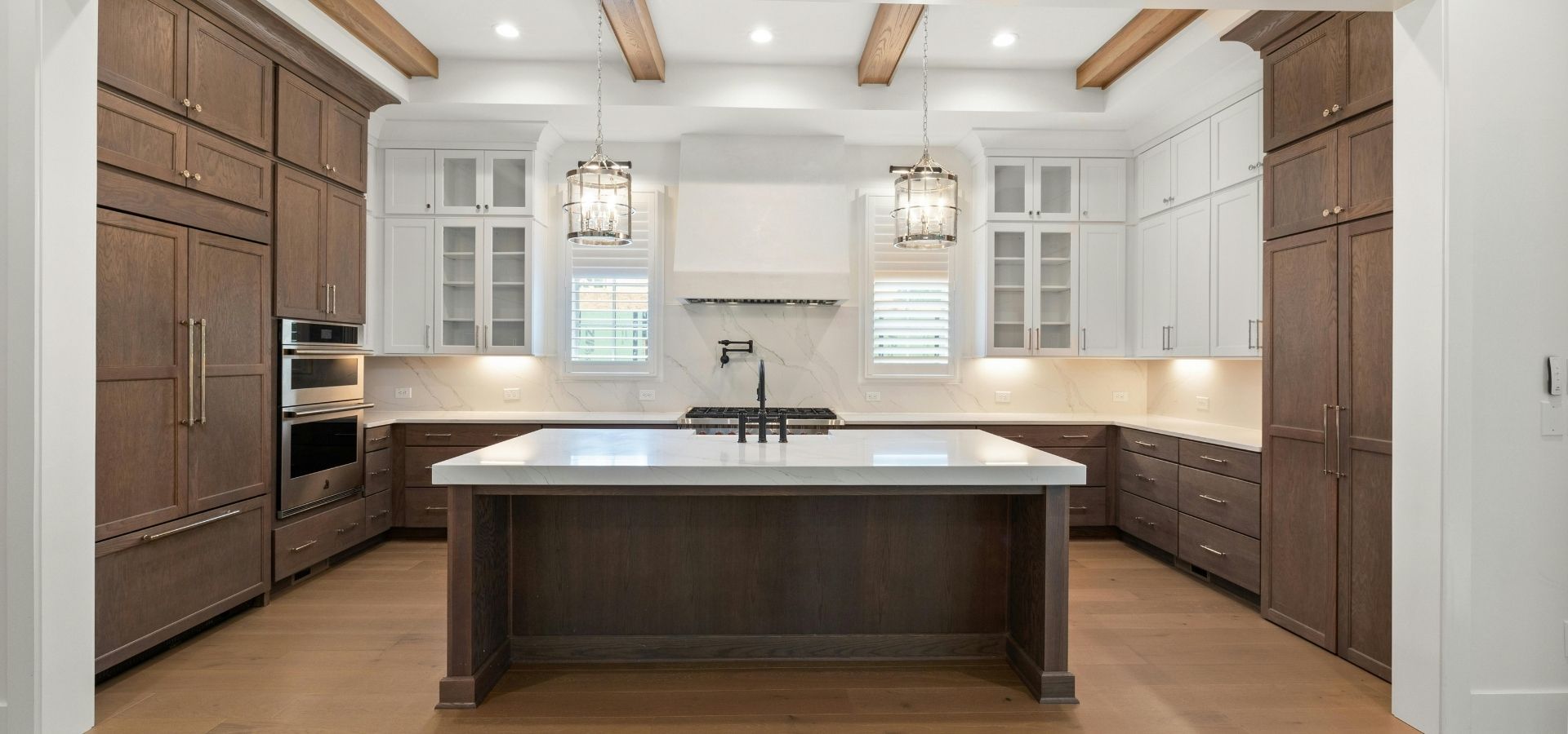 High-end custom kitchen featuring professional crown molding, dark wood cabinetry with white upper accents, and luxury ceiling beams over a massive marble waterfall island.