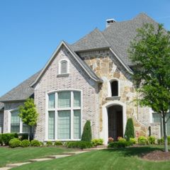 Luxury custom new construction home with a mix of light brick, natural stone accents, and high-pitched rooflines under a clear blue sky.