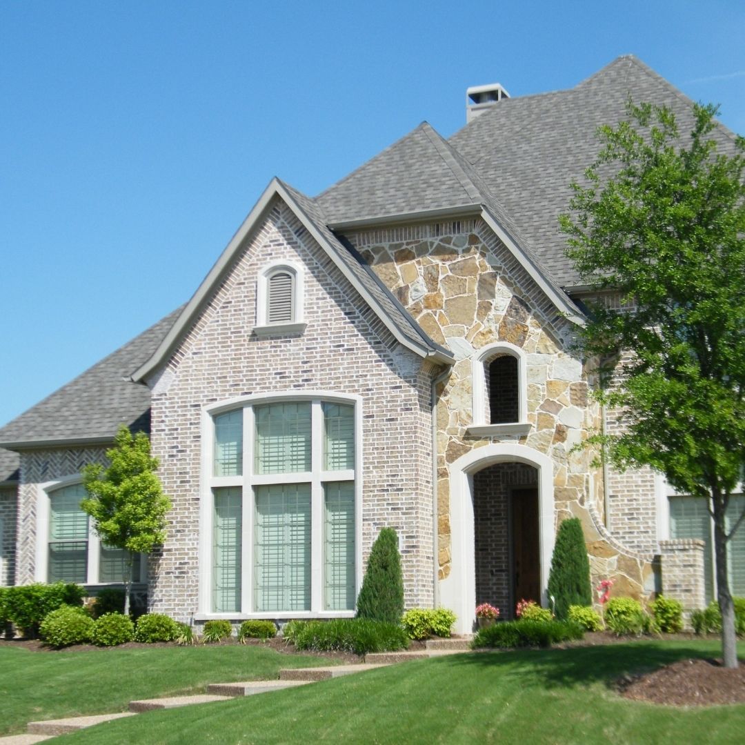 Luxury custom new construction home with a mix of light brick, natural stone accents, and high-pitched rooflines under a clear blue sky.