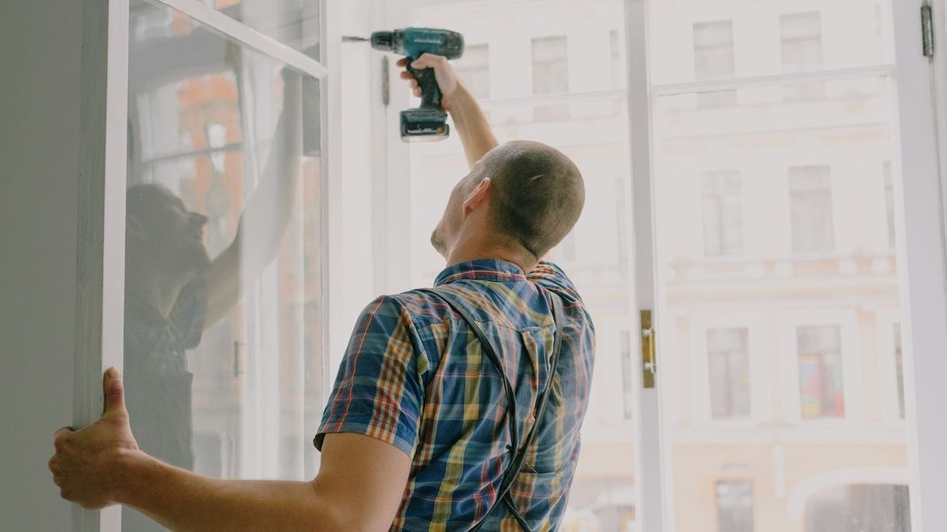 A professional installer using a power drill to secure a large window frame during a residential home improvement project.