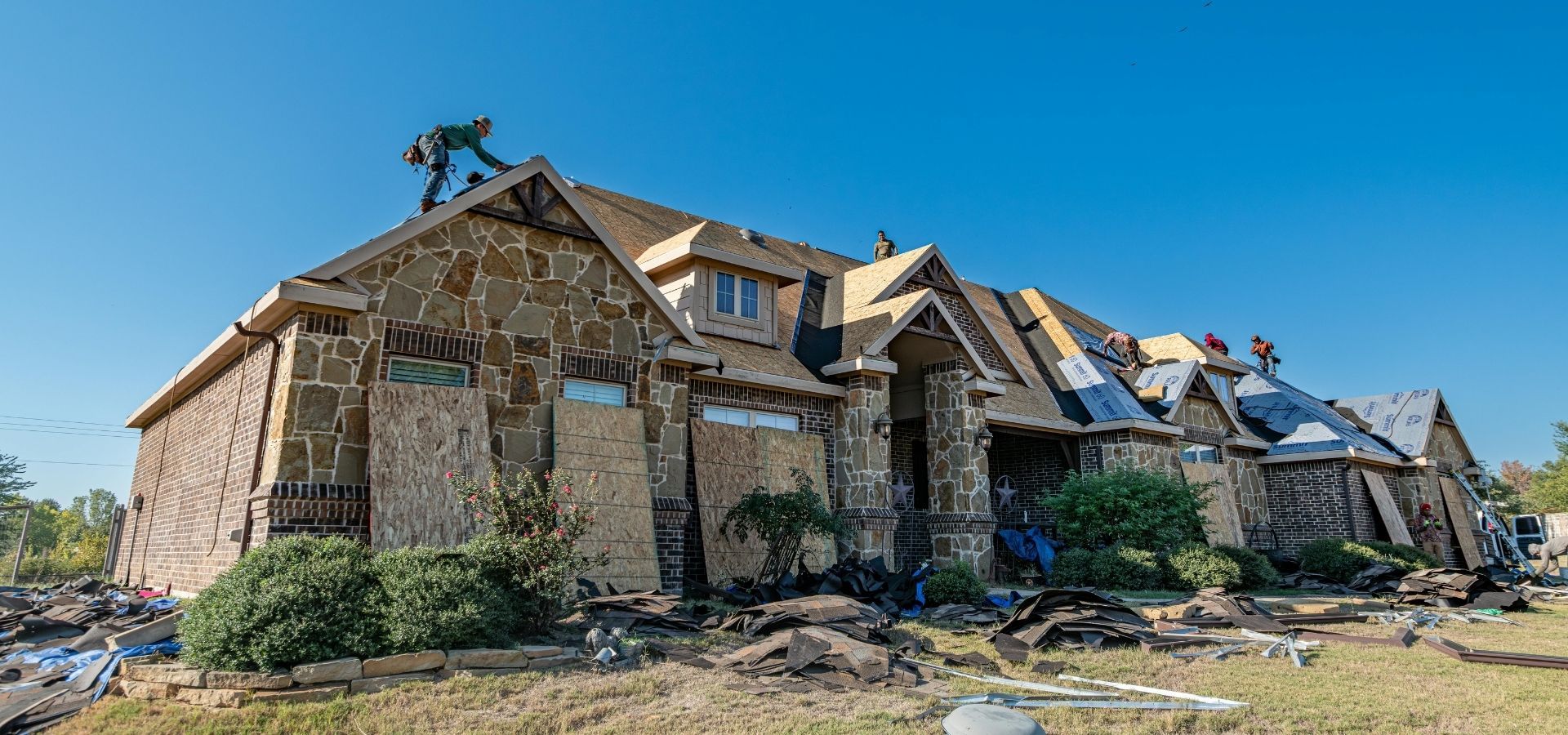 A team of professional roofers installing new asphalt shingles on a large, custom stone and brick residential home under a clear blue sky.