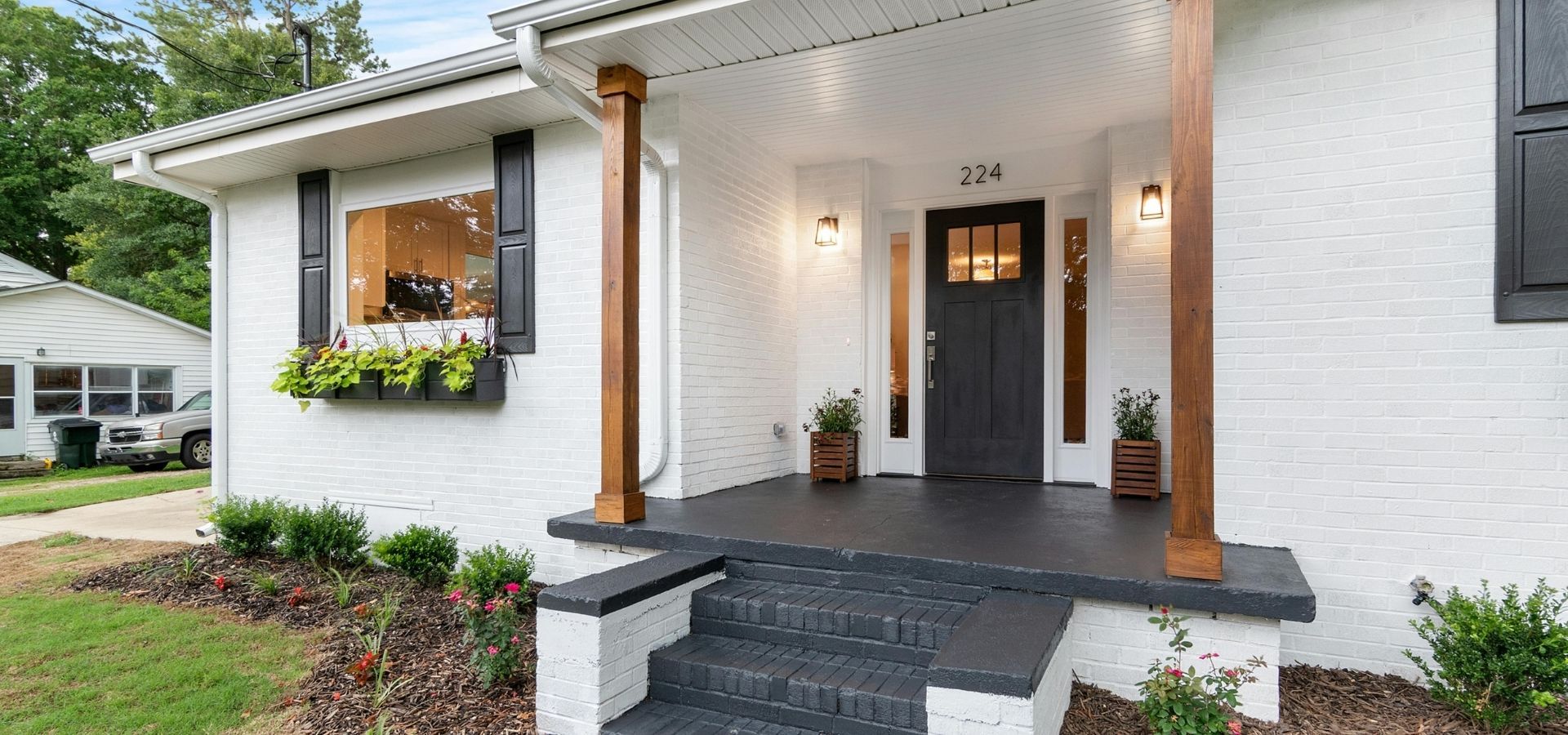 High-end curb appeal featuring a white brick home renovation with custom wood porch pillars, a black front door, and professional landscaping.