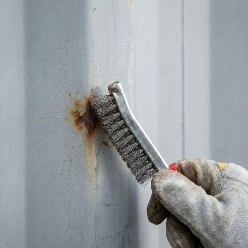 Gloved hand removing rust from a shipping container with a wire brush.