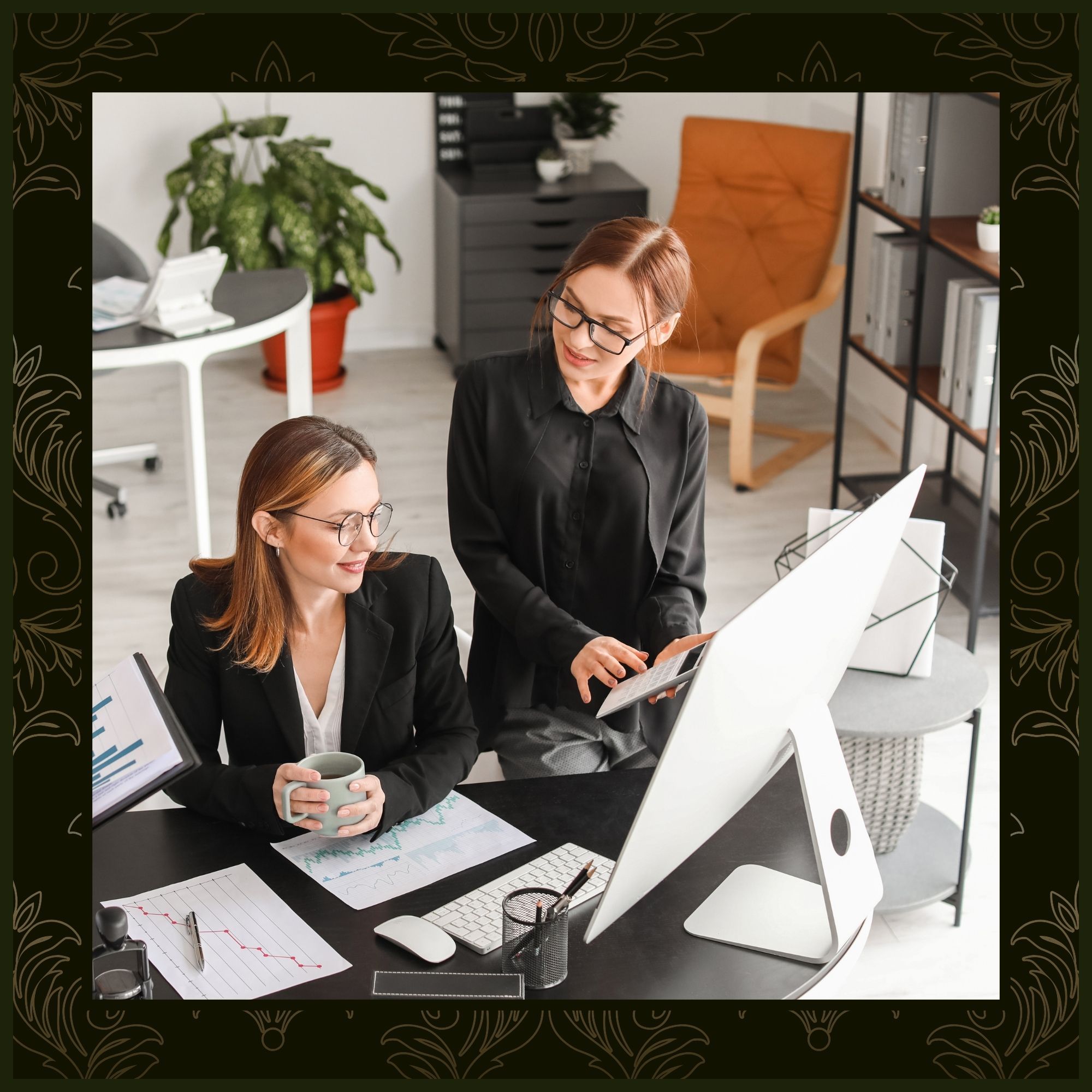 Two women in a modern office discuss data on a large computer monitor. One sits at the desk holding a mug, while the other stands beside her, pointing toward a tablet or calculator.