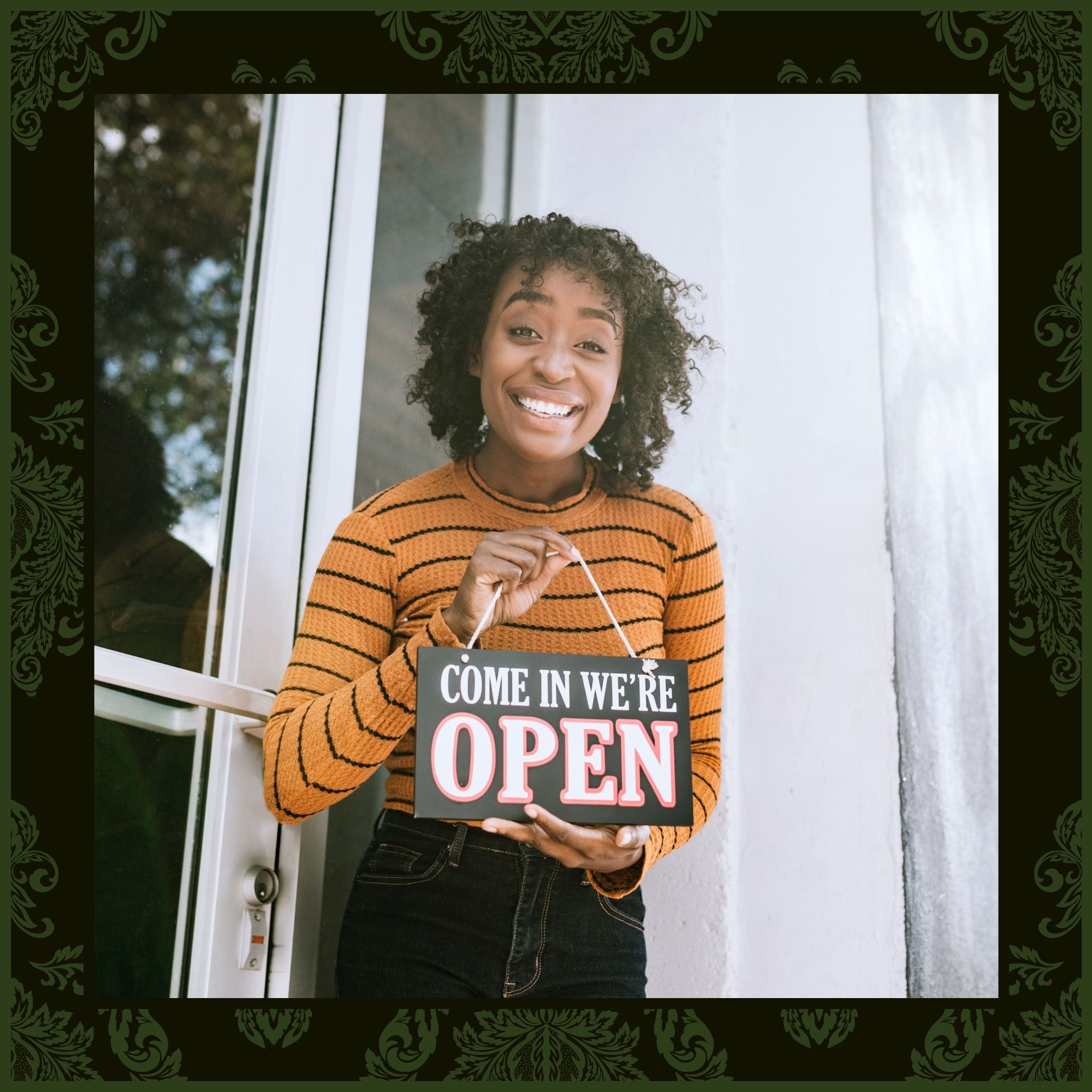 A smiling woman in a striped shirt holding a "Come In We’re Open" sign, framed by a dark green damask border.