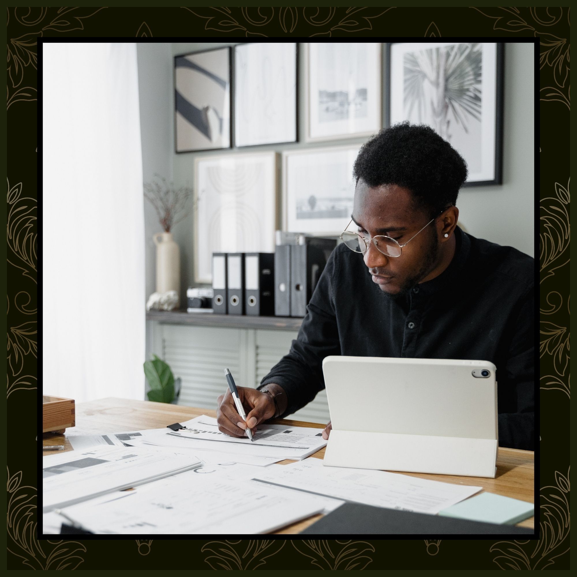 A man with glasses working at a wooden desk, writing on financial spreadsheets with a tablet propped open in front of him.