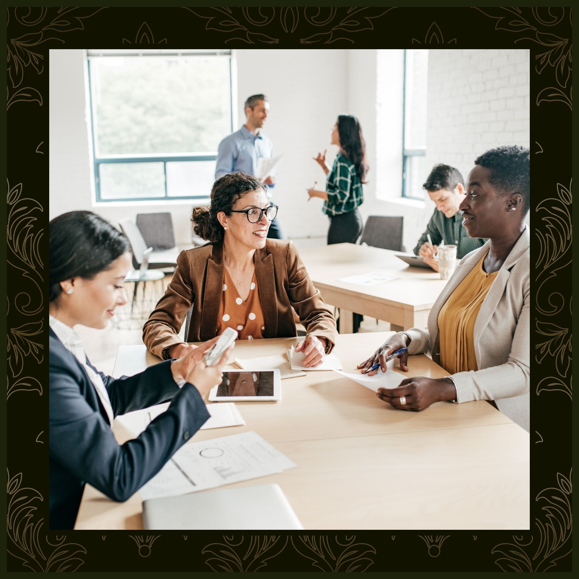 A diverse group of four professionals in business casual attire sit around a light wood conference table, engaged in a collaborative meeting. In the bright background, two other colleagues are seen standing and talking near a large window.