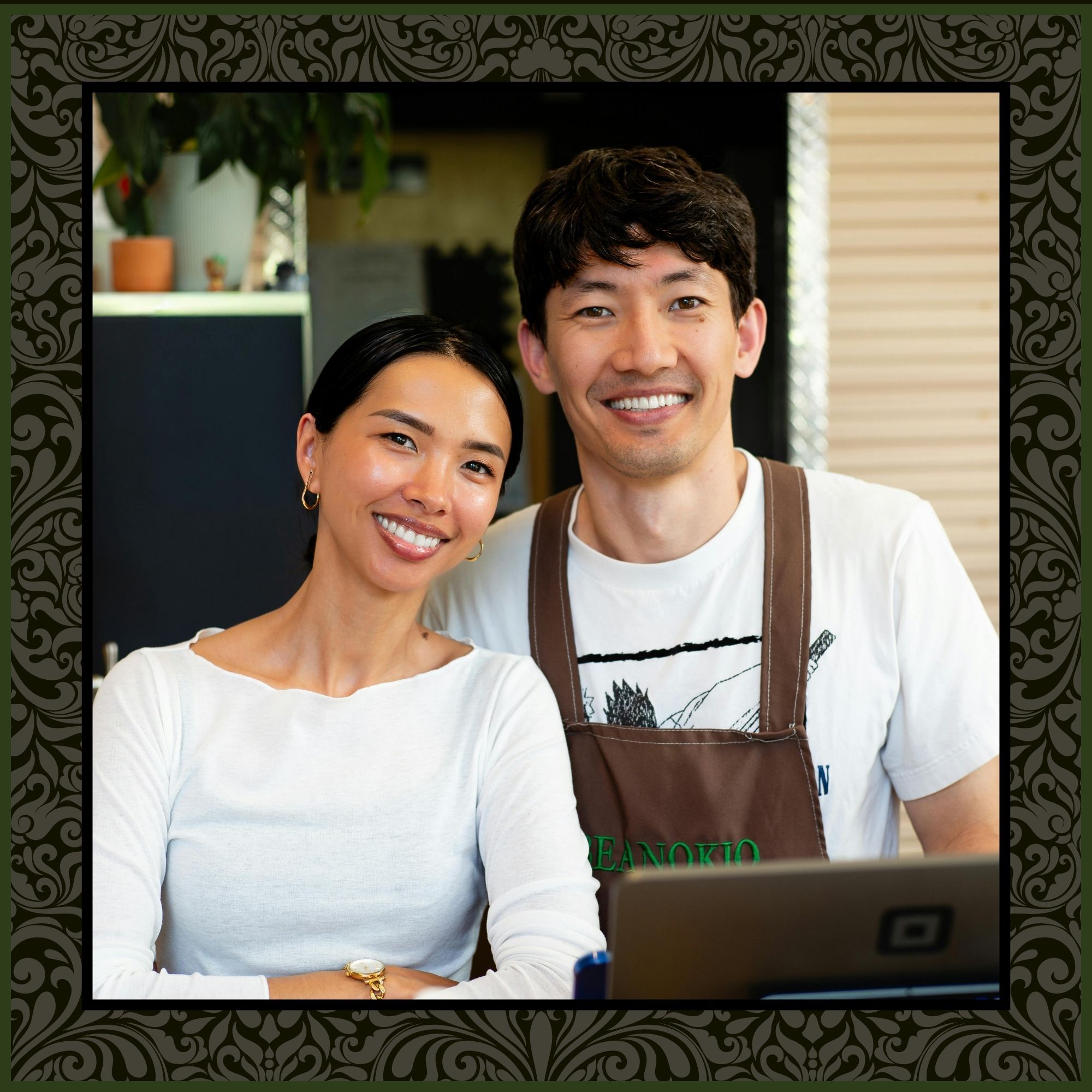 A friendly professional man and woman smiling behind a counter, framed by an intricate dark green scrollwork border.