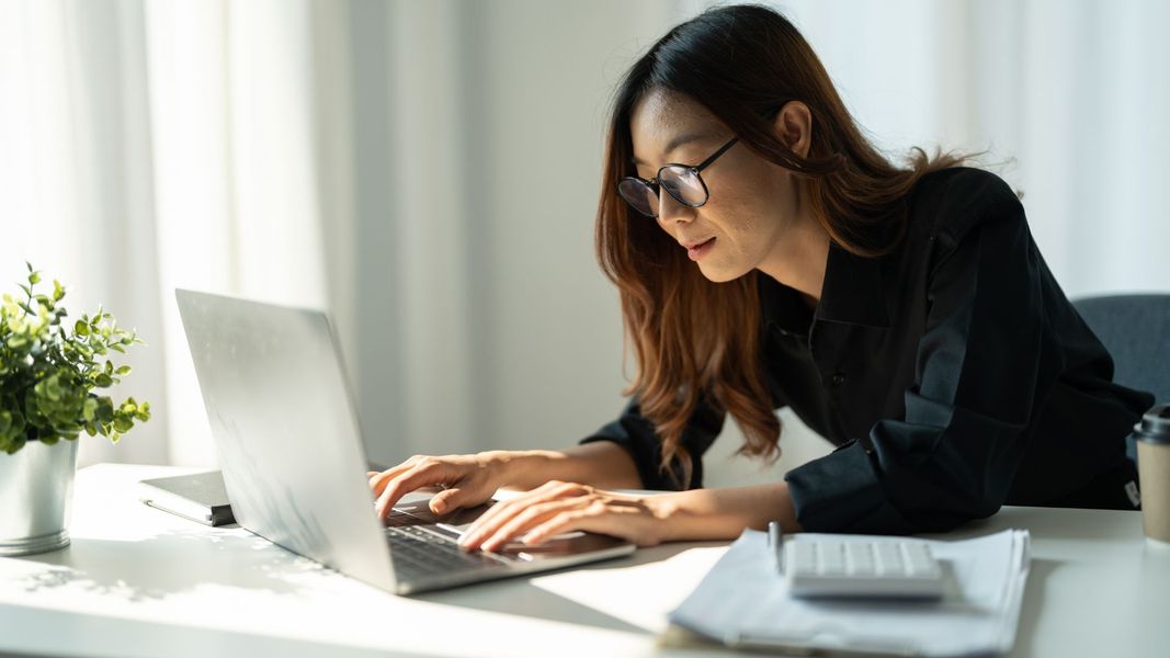 A professional woman with glasses and long brown hair working intently on a laptop at a bright, sunlit desk with a calculator and documents nearby. A professional woman with glasses and long brown hair working intently on a laptop at a bright, sunlit desk with a calculator and documents nearby.