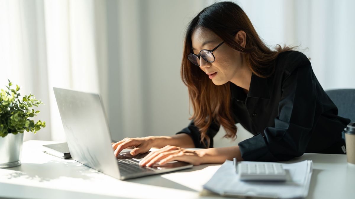 A professional woman with glasses and long brown hair working intently on a laptop at a bright, sunlit desk with a calculator and documents nearby.
