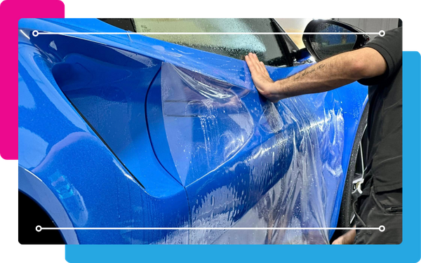 A hand smoothing a clear paint protection film over the fender of a vibrant blue car. The film is visible as a transparent layer being pressed down, highlighting the precise and professional application of auto paint protection film.