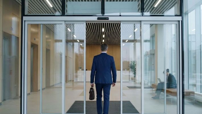 An automatic glass sliding door opens as a person in business attire enters a brightly lit, modern office lobby.