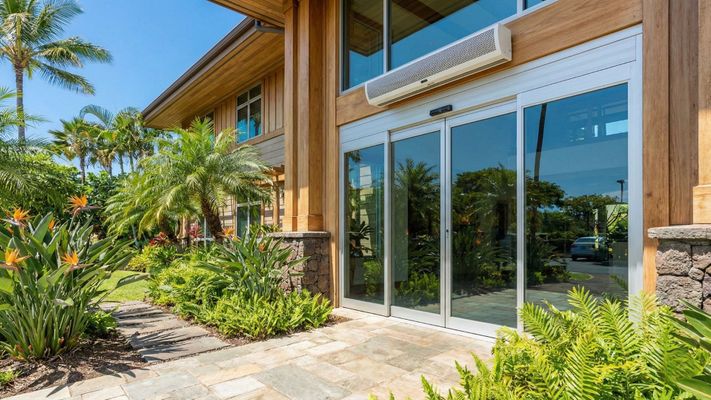 Modern Hawaii office building entrance with automatic sliding doors and an air curtain unit above, surrounded by tropical landscaping under bright sun.