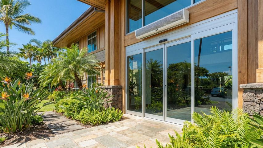 Modern Hawaii office building entrance with automatic sliding doors and an air curtain unit above, surrounded by tropical landscaping under bright sun.